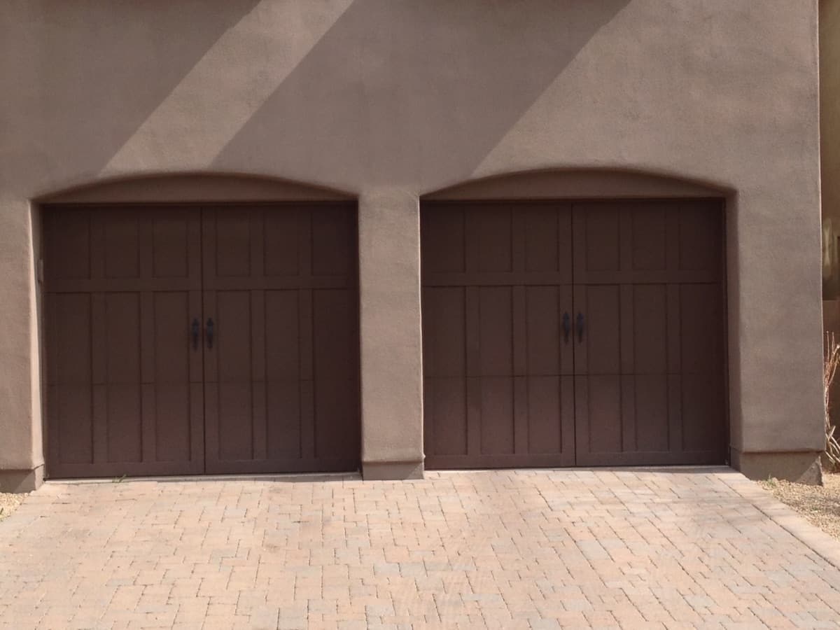 Two brown garage doors on a beige wall with a brick driveway