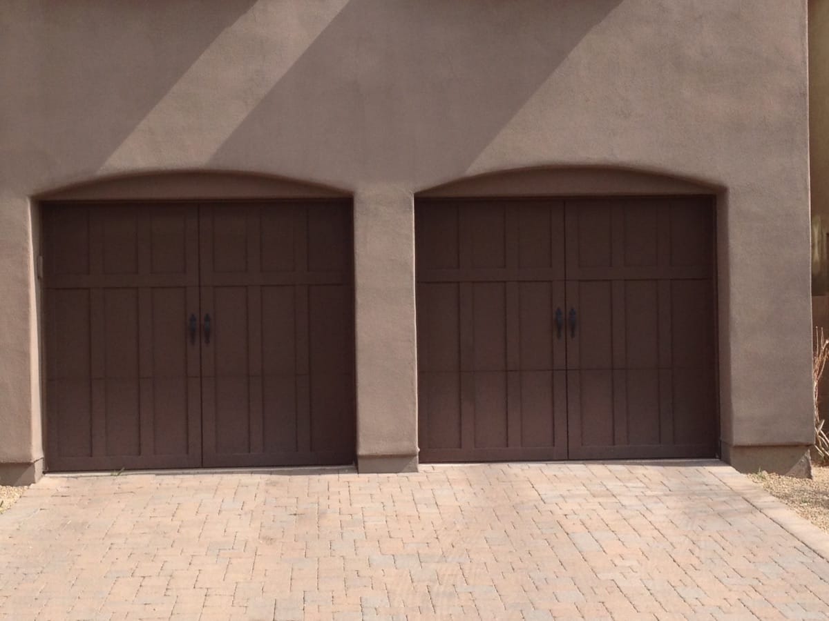 Two brown garage doors on a beige wall with a brick driveway